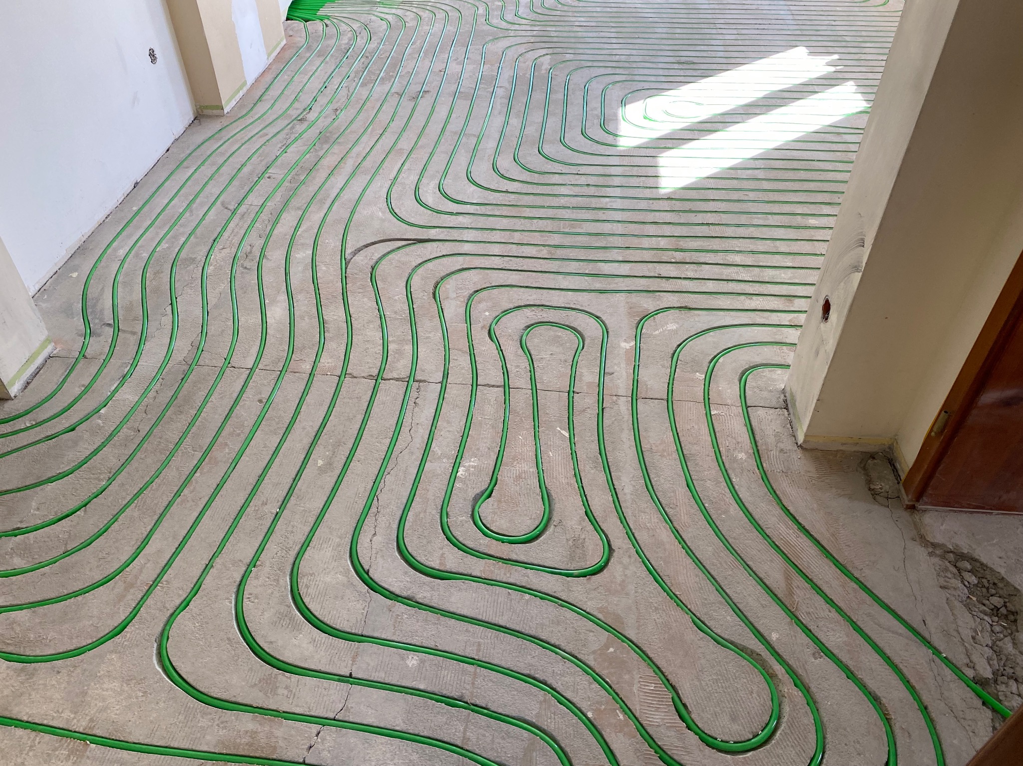 Zielone rurki ogrzewania podłogowego ułożone w pętle na szarej, surowej posadzce w jasnym pomieszczeniu. Widoczne promienie słoneczne.