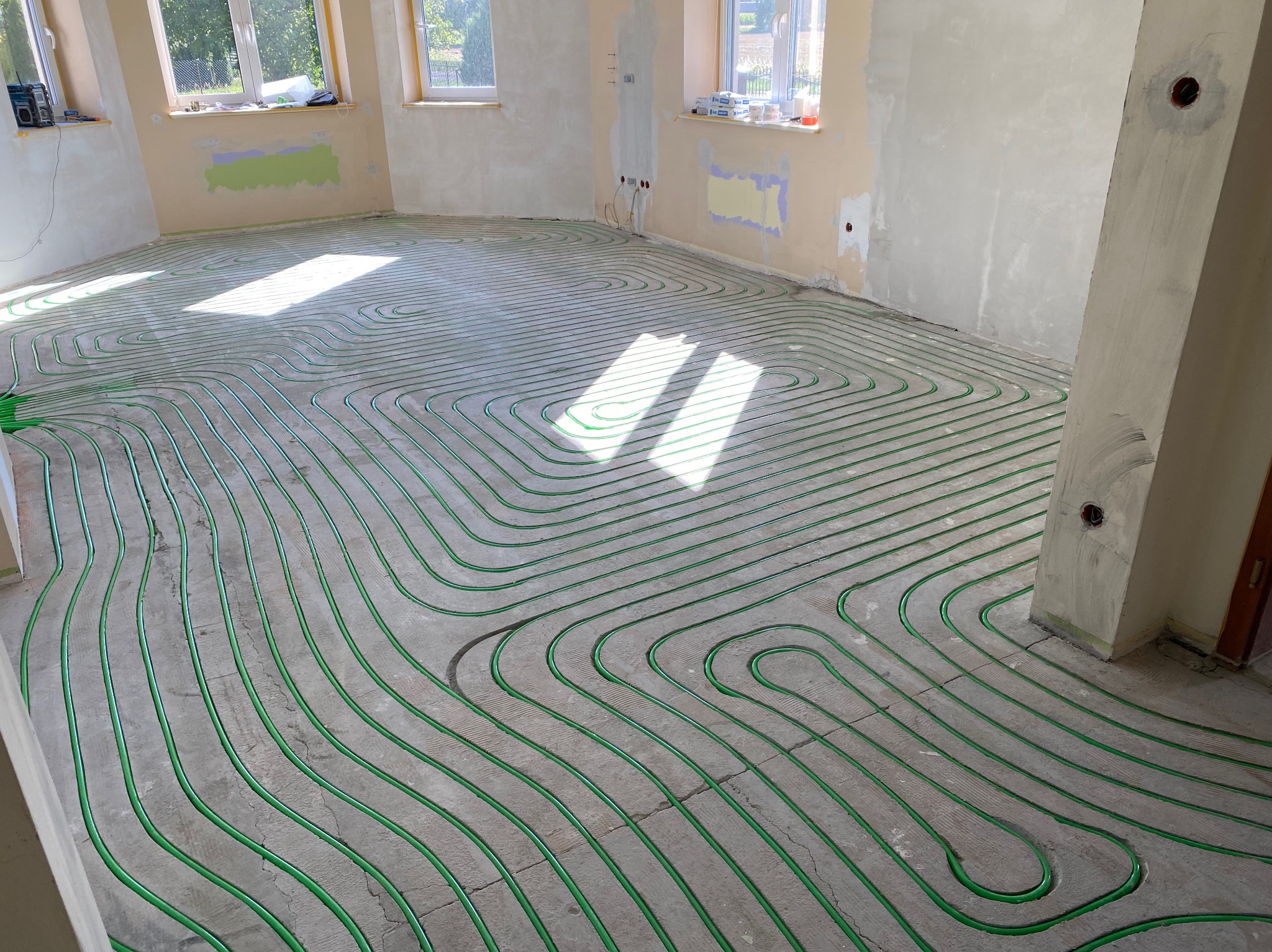 Zielone rury ogrzewania podłogowego ułożone w jodełkę na szarej, betonowej posadzce w jasnym pomieszczeniu z oknami. Widoczne ślady po pracach wykończeniowych.