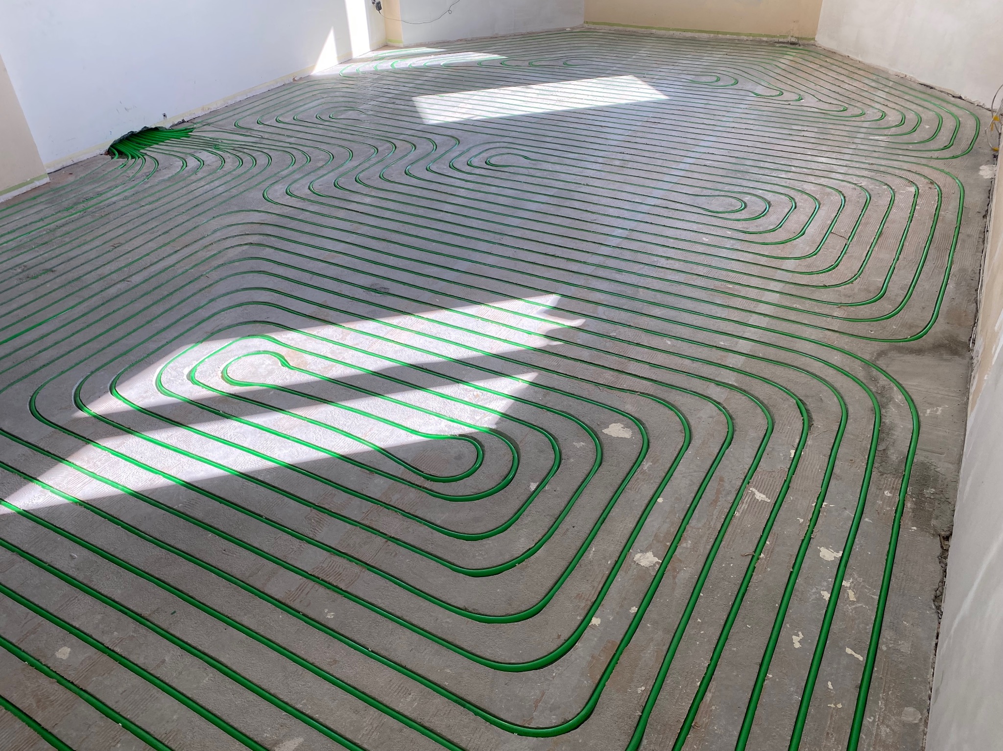 Zielone rurki ogrzewania podłogowego ułożone w pętle na szarej, surowej posadzce w jasnym pomieszczeniu. Widoczne promienie słoneczne.