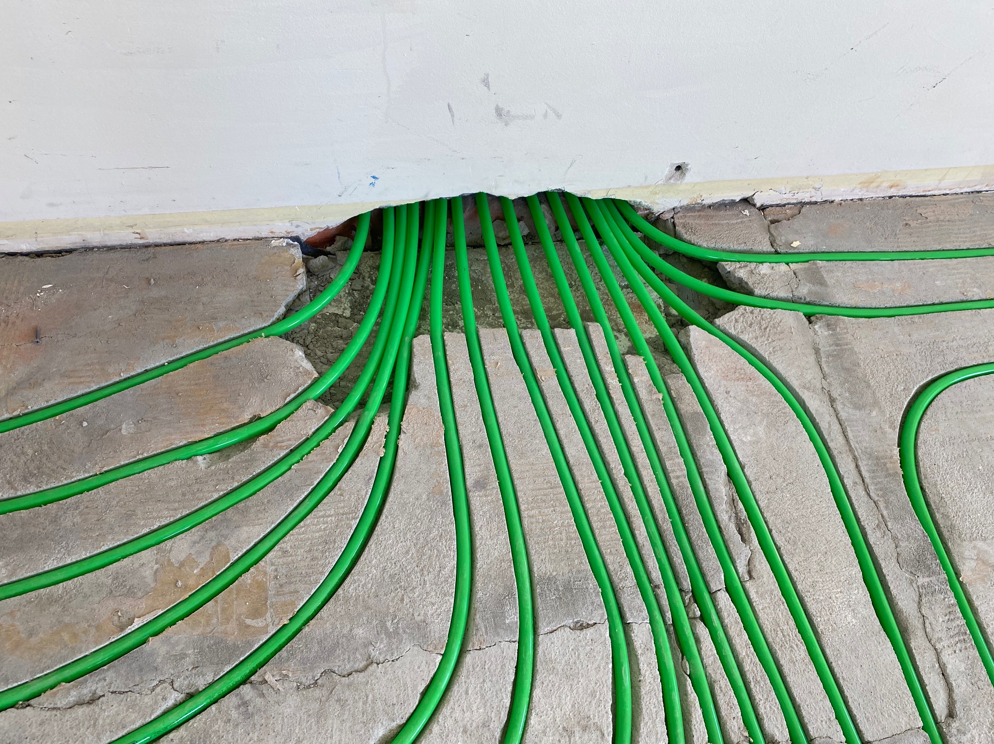 Zielone rurki ogrzewania podłogowego wychodzące ze ściany na surową, betonową podłogę. Widoczne pęknięcia i nierówności podłoża. Instalacja w trakcie realizacji.