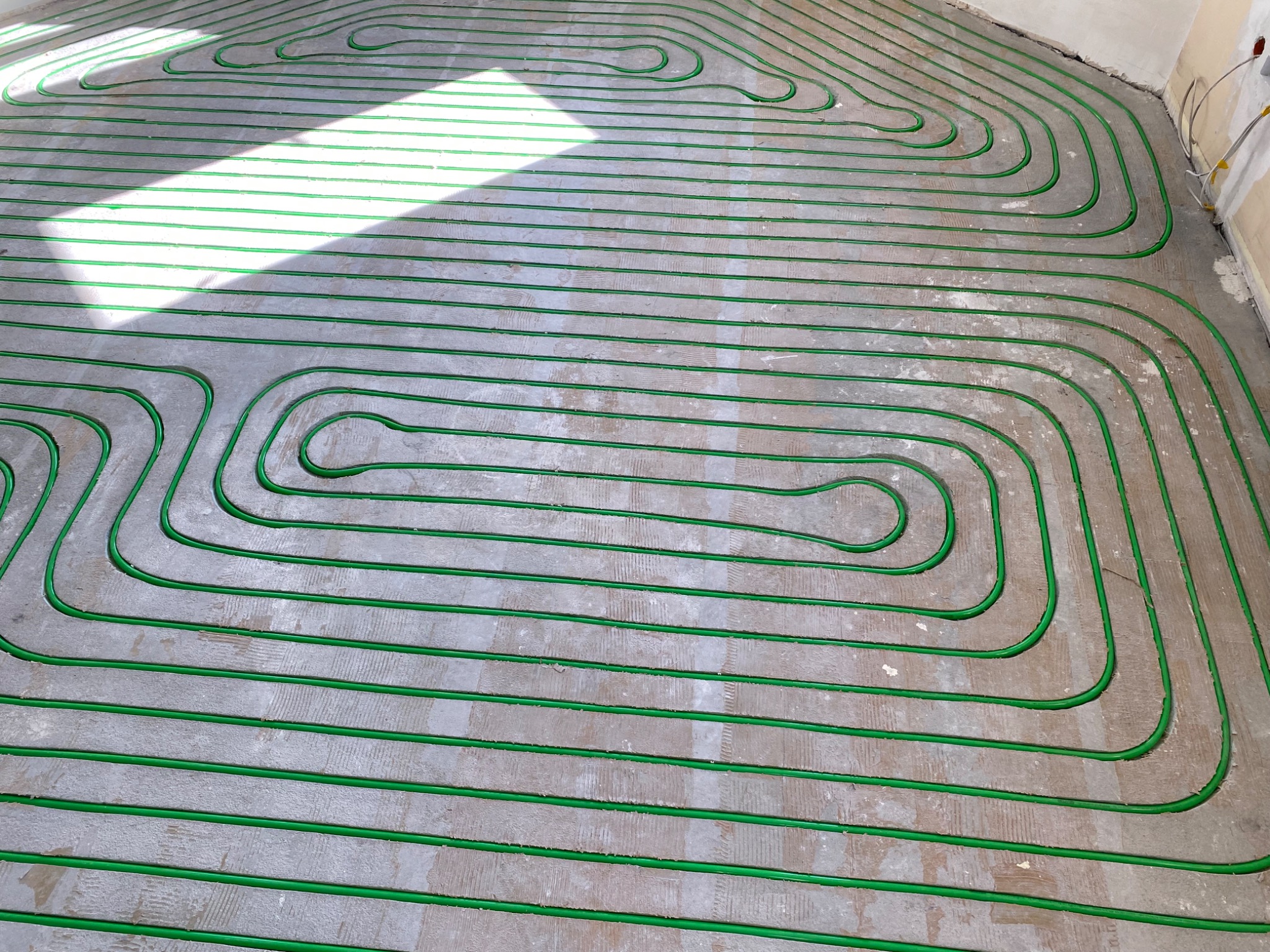 Zielone rurki ogrzewania podłogowego ułożone spiralnie na szarej, betonowej posadzce w jasnym pomieszczeniu. Widoczny fragment ściany i padające światło słoneczne.