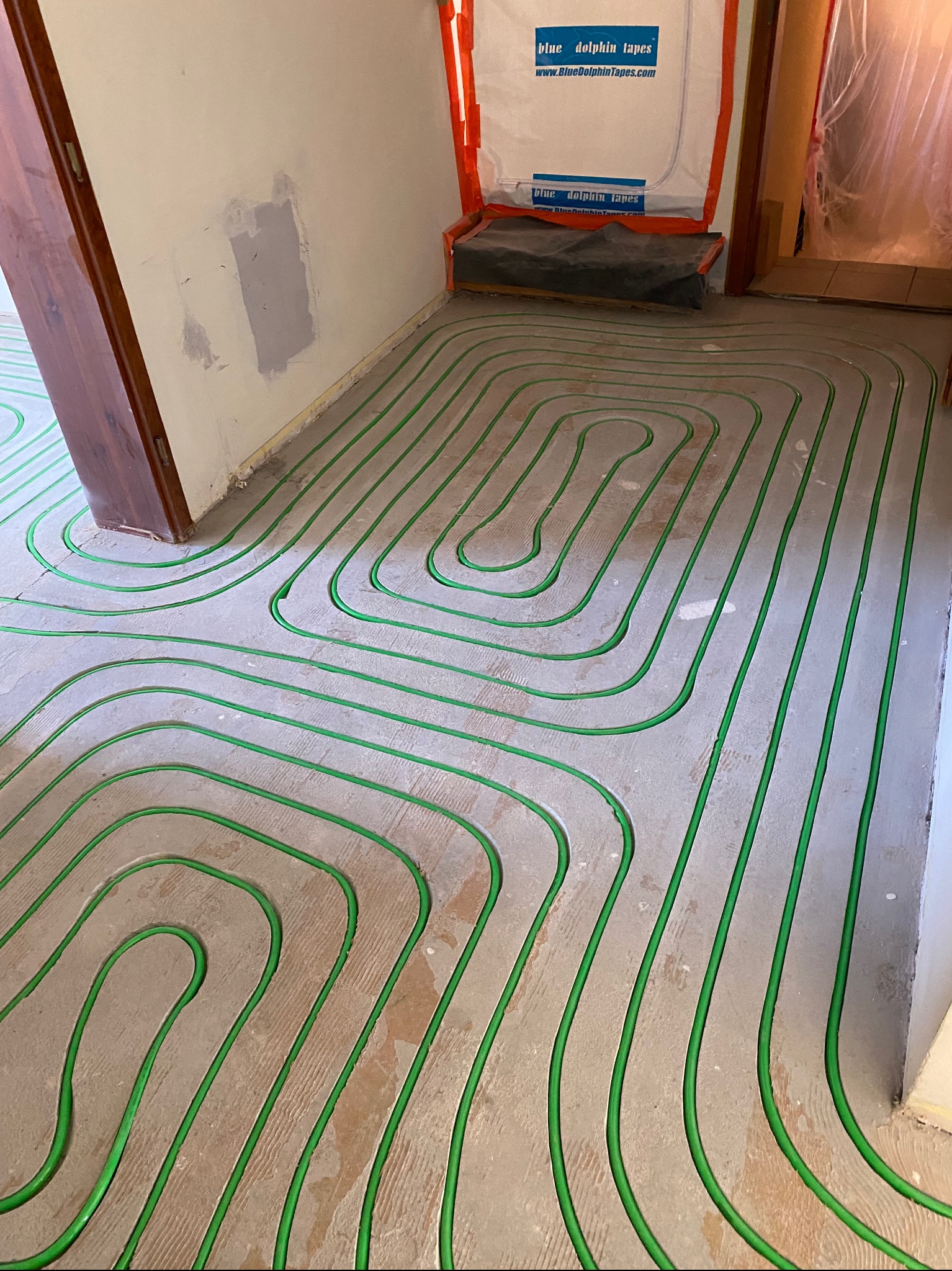 Zielone rurki ogrzewania podłogowego ułożone spiralnie na surowej posadzce, widok z góry. Fragment ściany z widocznymi śladami po naprawach.