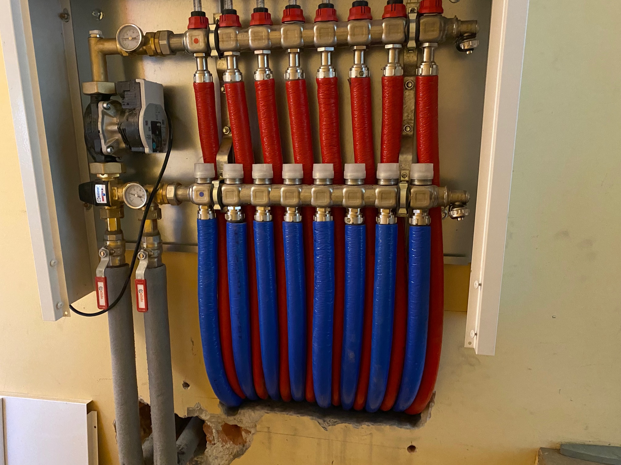 Rozdzielacz ogrzewania podłogowego z czerwonymi i niebieskimi rurami, zamontowany na ścianie. Widoczne zawory i manometry. Instalacja w trakcie montażu, z fragmentem ściany do wykończenia.