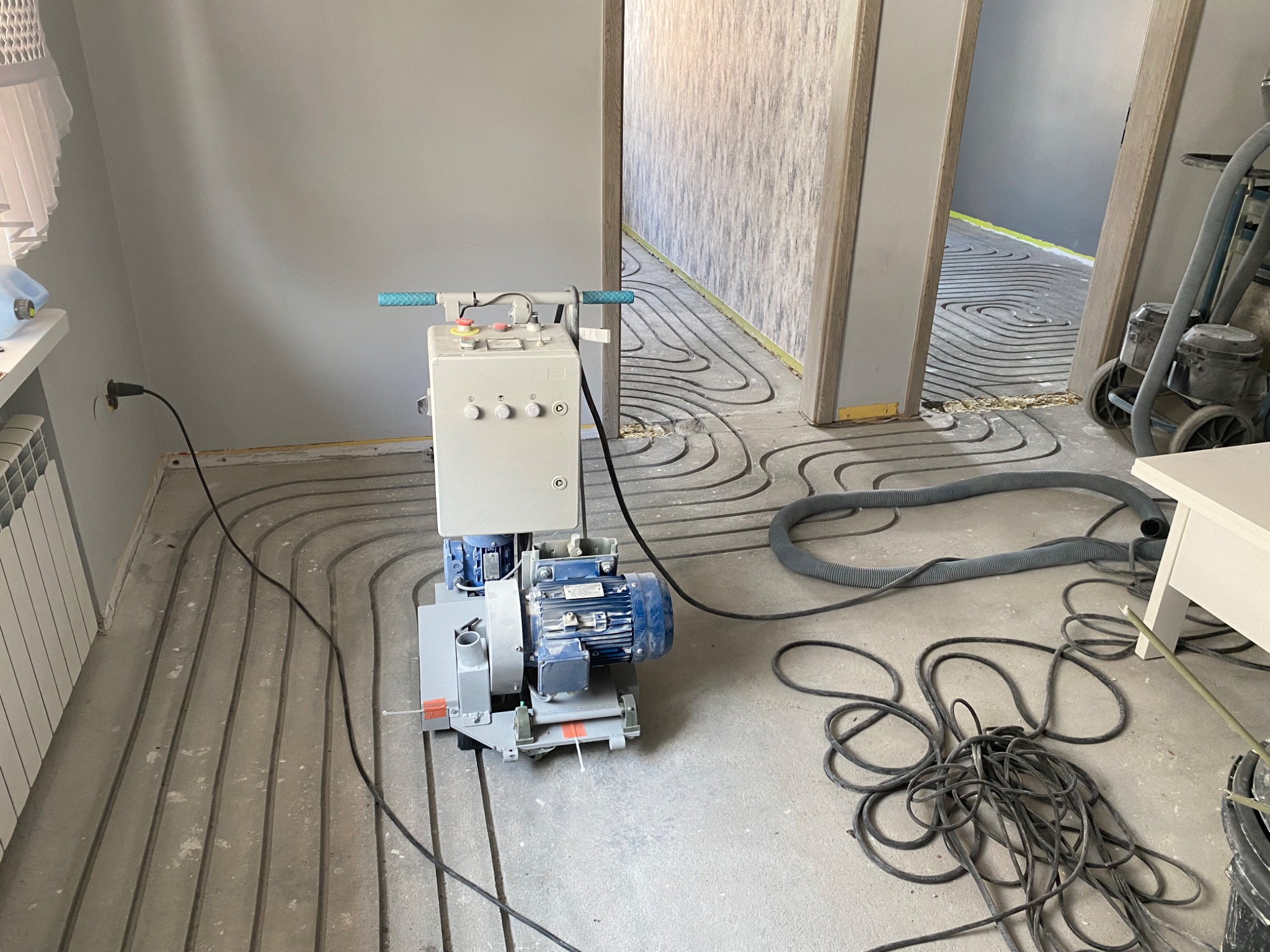 Szlifowanie wylewki pod ogrzewanie podłogowe maszyną z niebieskim silnikiem. Widoczne rury instalacji grzewczej w szlifowanej posadzce. Szare ściany i bałagan.