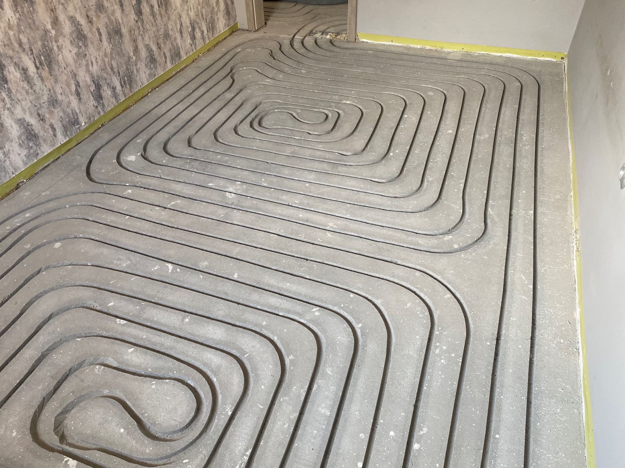 Szare rury ogrzewania podłogowego ułożone spiralnie w pomieszczeniu w Mikstacie, przygotowane do zalania wylewką. Widoczne fragmenty ścian i żółta taśma dylatacyjna.