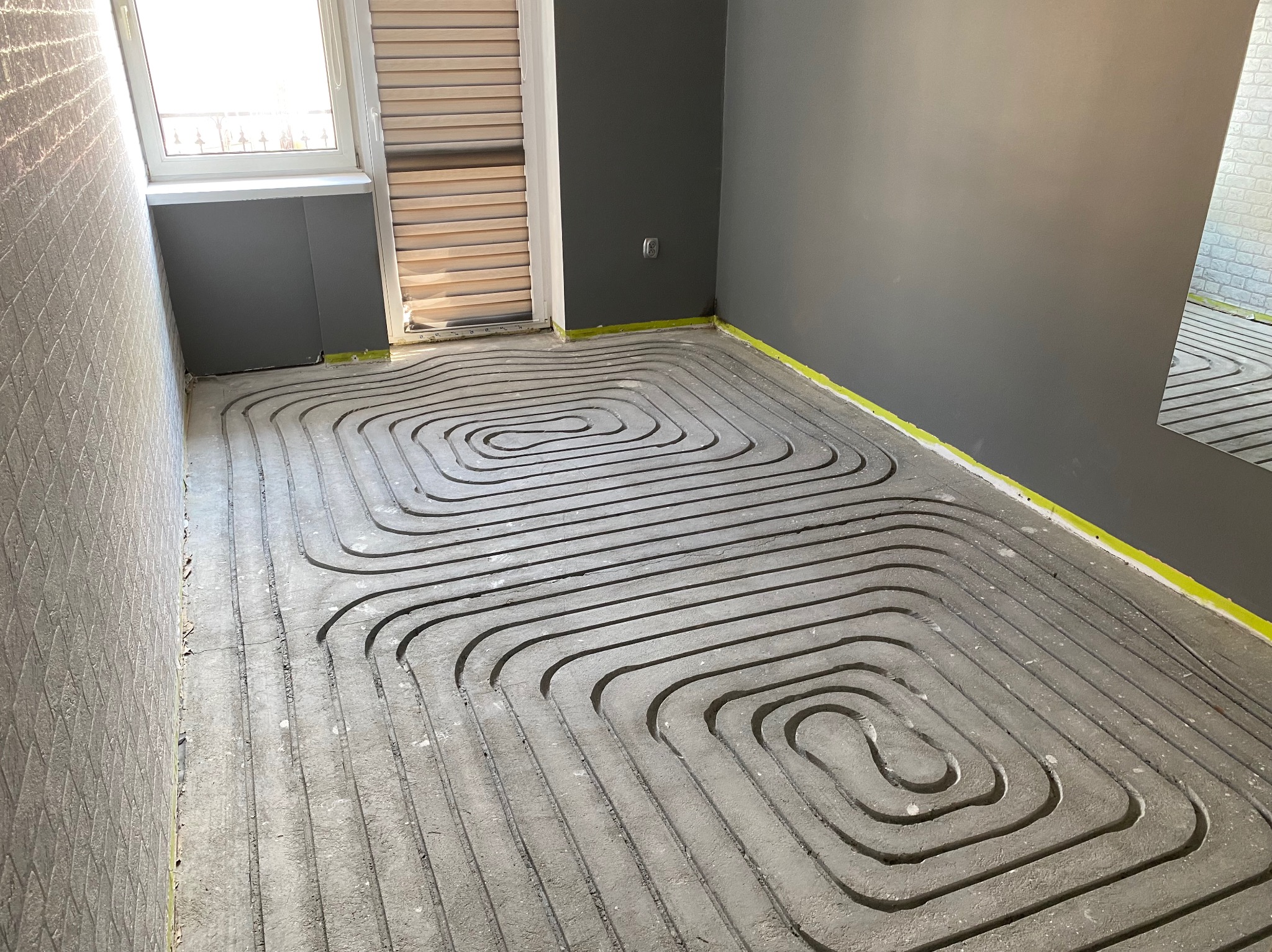 Szare rury ogrzewania podłogowego ułożone spiralnie w pokoju z oknem i szarą ścianą. Żółta taśma na styku podłogi i ściany. Widok z góry na instalację.