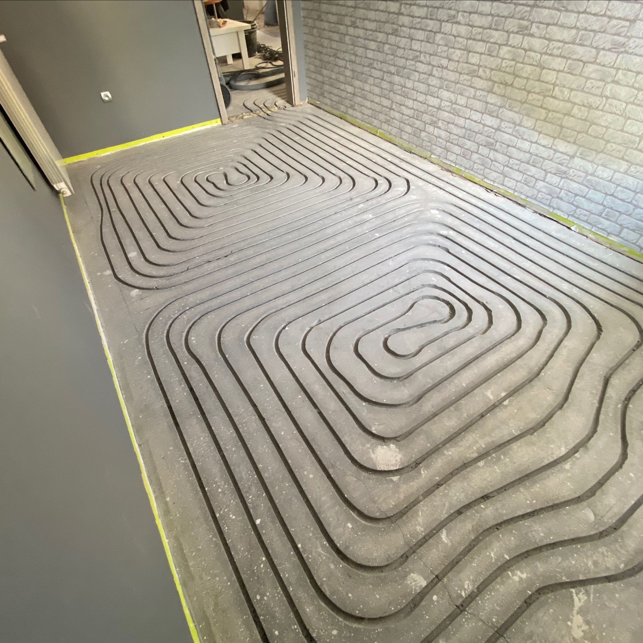 Szare rury ogrzewania podłogowego ułożone spiralnie na przygotowanym podłożu, widoczne taśmy zabezpieczające przy ścianach i fragment ściany z imitacją cegły.