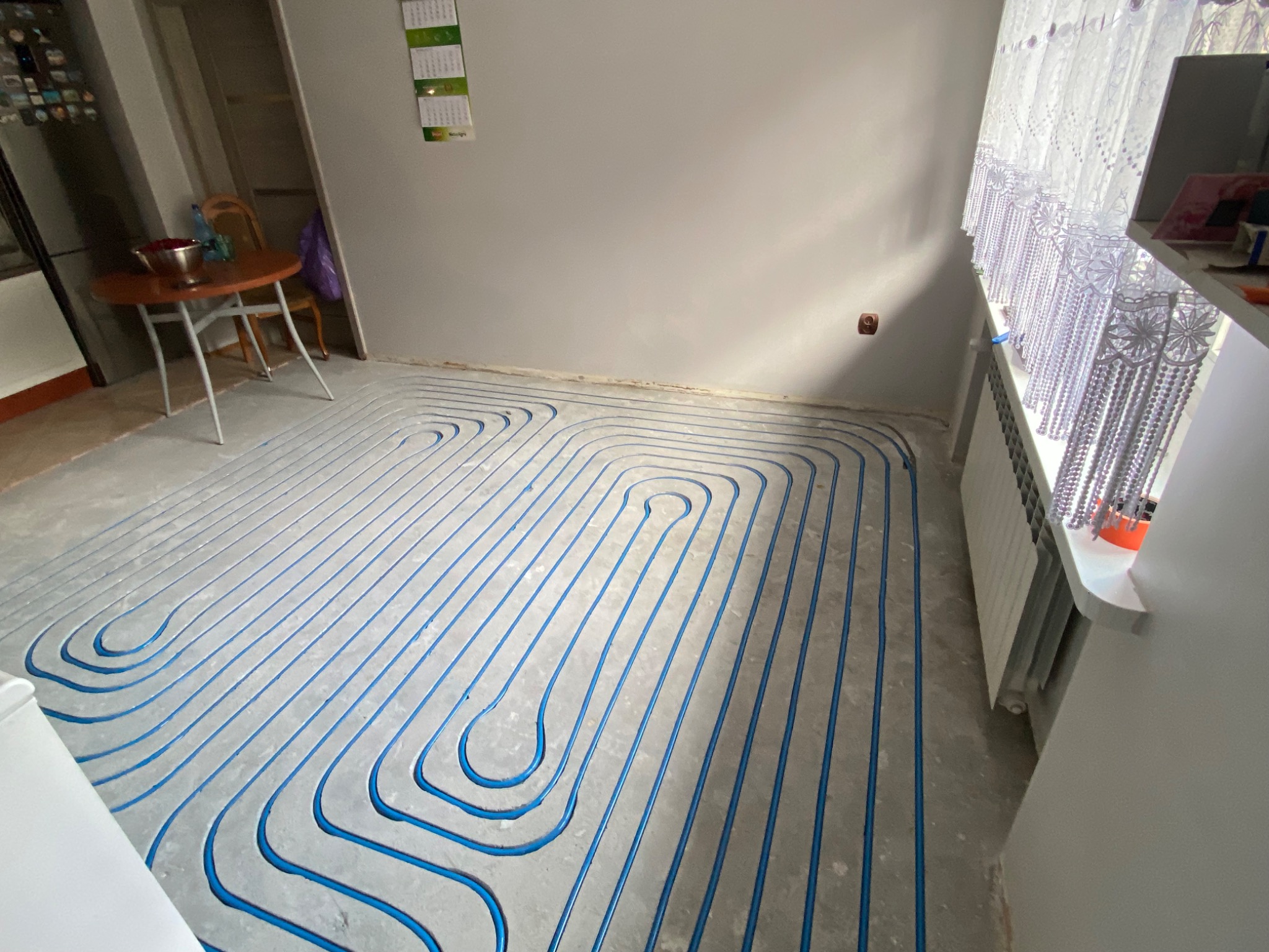 Niebieskie rurki ogrzewania podłogowego ułożone w pętle na szarej wylewce, gotowe do zalania. Widok z góry, fragment okna z firanką w tle.