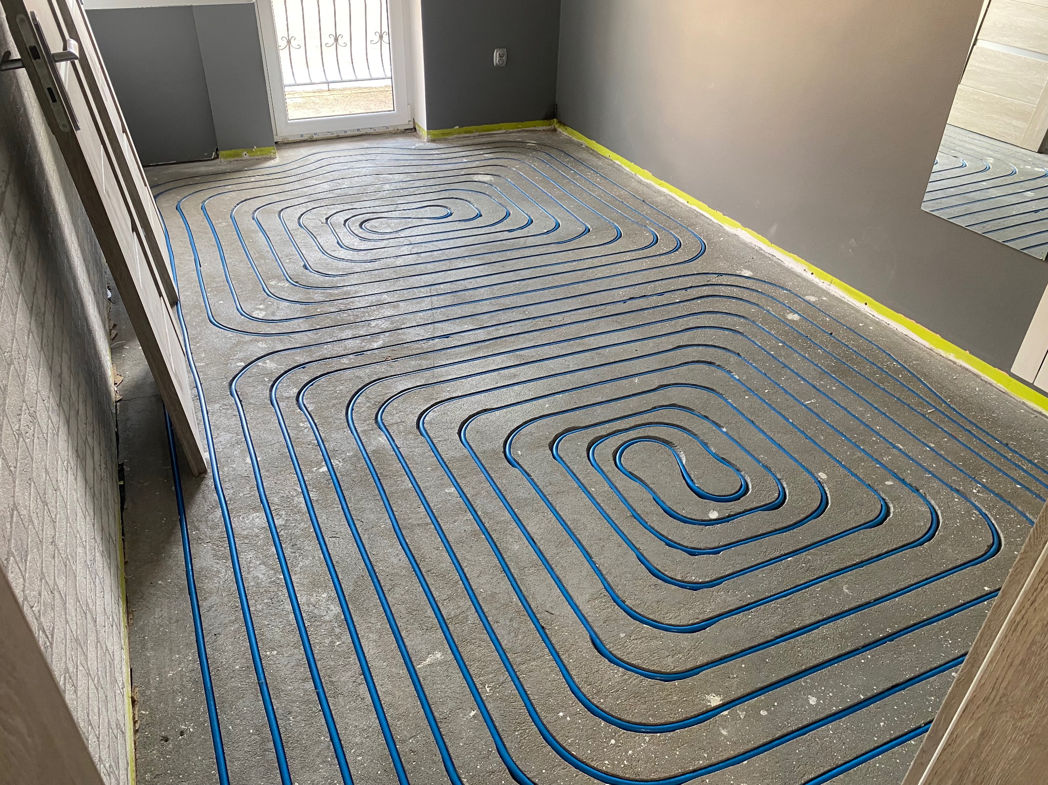 Niebieskie rurki ogrzewania podłogowego ułożone spiralnie na szarym betonie, widoczne żółte taśmy przy ścianach i drabina oparta o ścianę.