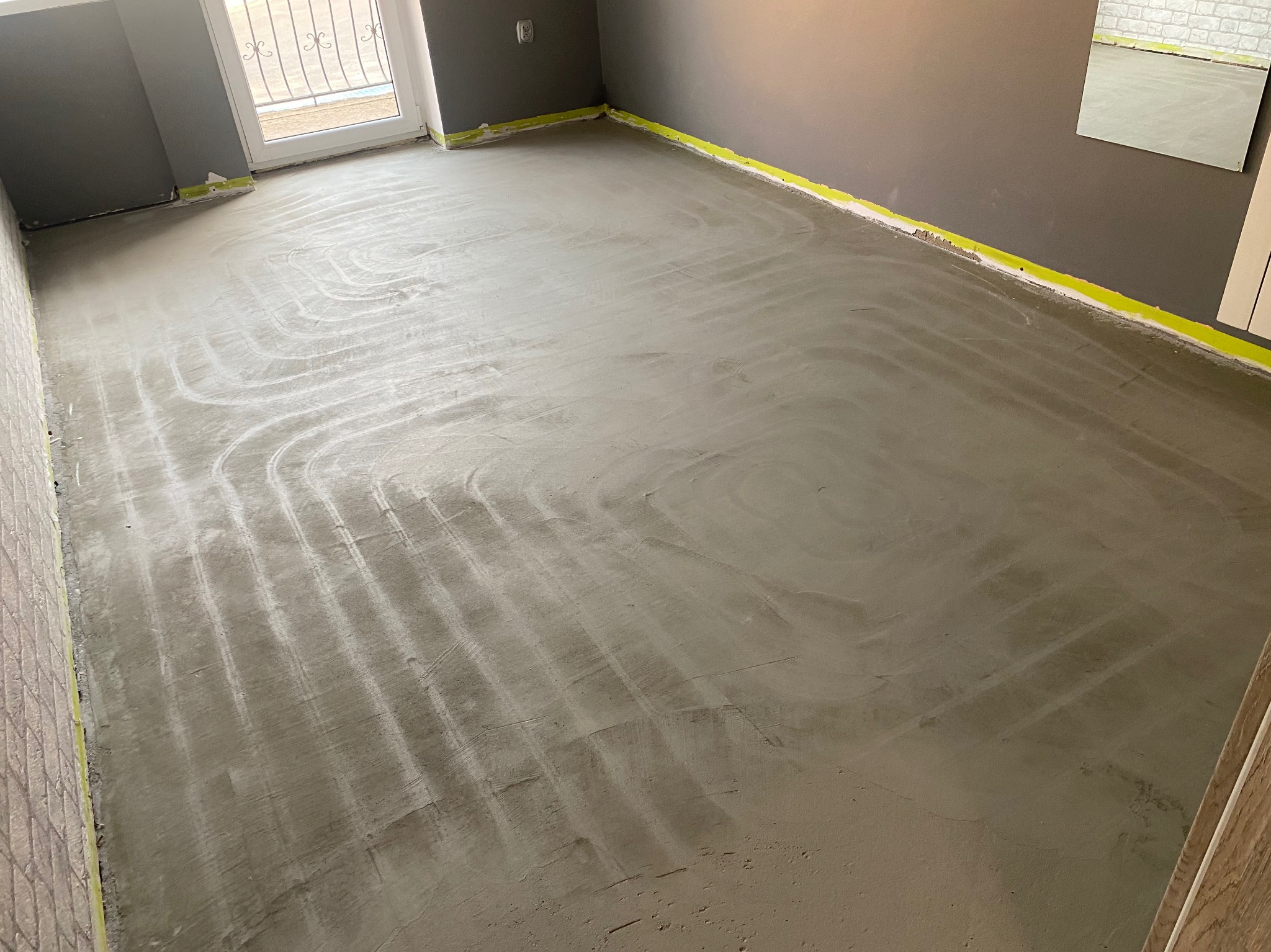 Wylana, szara posadzka z widocznymi śladami po instalacji ogrzewania podłogowego. Żółta taśma maskująca przy ścianach. Fragment okna i drzwi balkonowych w tle.