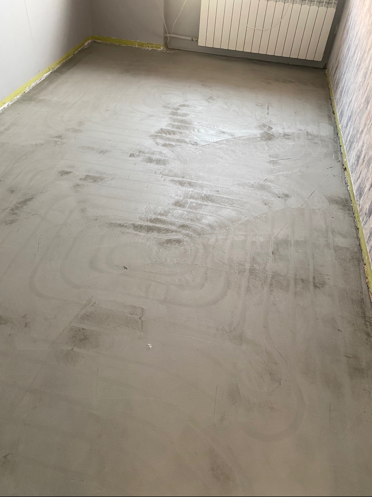 Wylana, świeża wylewka podłogowa w pomieszczeniu, zabezpieczona taśmą malarską przy ścianach. Widoczny grzejnik na ścianie i ślady po rozprowadzaniu masy.