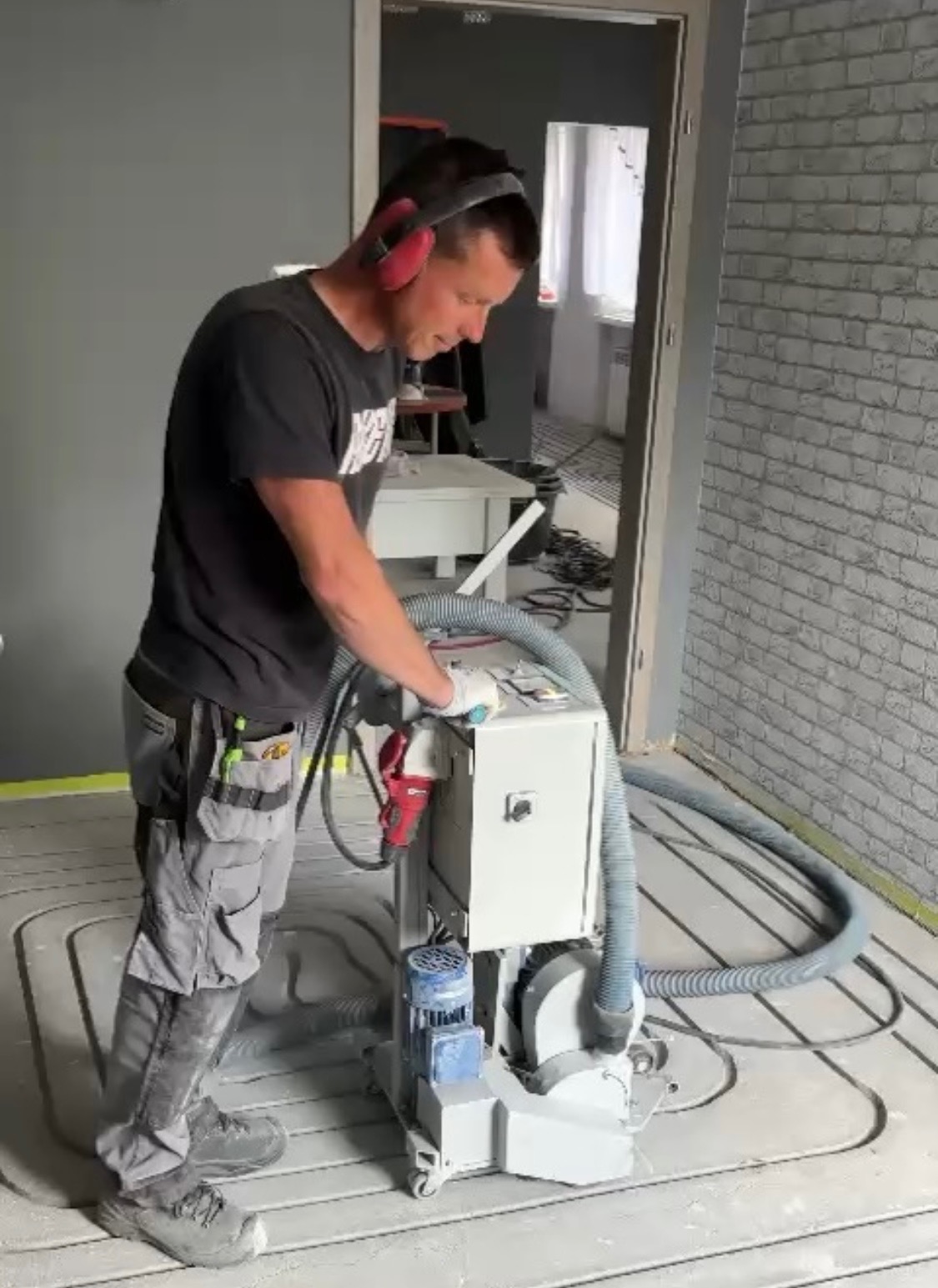 Pracownik w słuchawkach szlifuje posadzkę z ogrzewaniem podłogowym za pomocą specjalistycznej maszyny. Widoczne rury ogrzewania w podłodze i ściana z cegły.