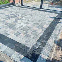 Ukończona nawierzchnia z szarej kostki brukowej o nieregularnym kształcie, obramowana pasem ciemnoszarej kostki, widoczna brama przesuwna i fragment trawnika w tle.