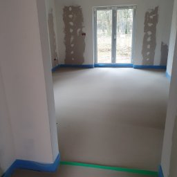 TB Tania Budowa - Wnętrze w stanie deweloperskim z widocznymi śladami po tynkowaniu ścian, zabezpieczoną folią podłogą i oknem balkonowym na wprost.