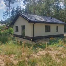 TB Tania Budowa - Niewielki parterowy dom z jasną elewacją i ciemnym dachem, otoczony wysoką trawą i drzewami, widoczny niebieski wąż ogrodowy.