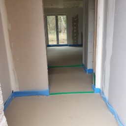 TB Tania Budowa - Wnętrze domu w trakcie prac wykończeniowych: wylewka podłogowa zabezpieczona niebieską pianką, ściany otynkowane, widoczne przygotowania do dalszych prac budowlanych.