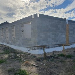 TB Tania Budowa - Wznoszone ściany domu z betonu komórkowego na fundamencie z izolacją, widoczne zbrojenia, palety i fragment dachu sąsiedniego budynku.