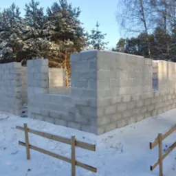 Wznoszenie ścian z bloczków betonowych w zimowej scenerii, widoczne fragmenty ścian z otworami okiennymi, ośnieżone podłoże i las w tle.