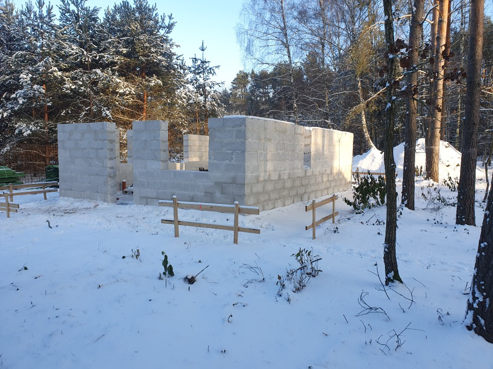 Wczesny etap budowy domu z bloczków betonowych w zimowej scenerii, widoczne fundamenty i pierwsze ściany, drewniane barierki zabezpieczające teren budowy, śnieg pokrywa ziemię i drzewa w tle.
