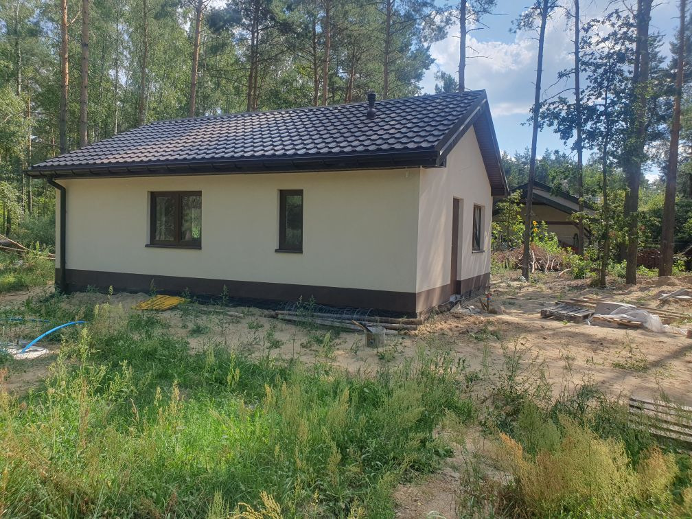 Gotowy, parterowy dom murowany z brązowymi oknami i ciemnobrązowym dachem, otoczony wysoką trawą i drzewami.