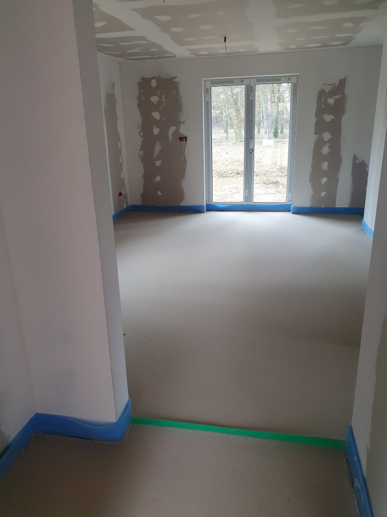 Wnętrze w stanie deweloperskim z widocznymi śladami po tynkowaniu ścian, zabezpieczoną folią podłogą i oknem balkonowym na wprost.