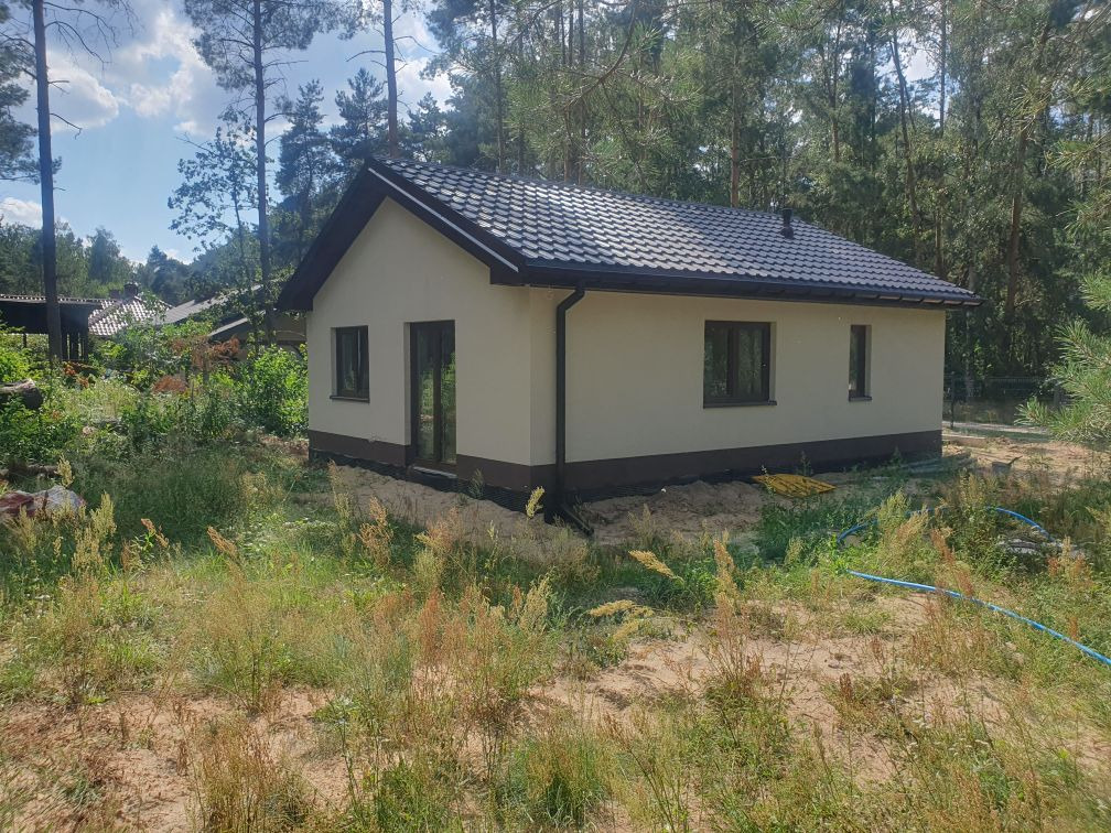 Niewielki parterowy dom z jasną elewacją i ciemnym dachem, otoczony wysoką trawą i drzewami, widoczny niebieski wąż ogrodowy.