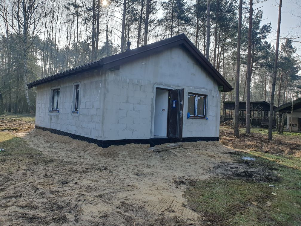 Surowy stan domu z bloczków, z otwartymi drzwiami wejściowymi i zamontowanymi oknami, widoczna izolacja fundamentów i piaszczyste podłoże.