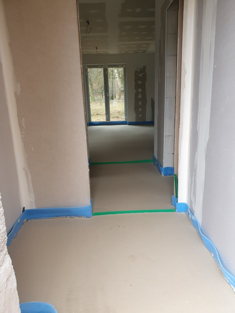 Wnętrze domu w trakcie prac wykończeniowych: wylewka podłogowa zabezpieczona niebieską pianką, ściany otynkowane, widoczne przygotowania do dalszych prac budowlanych.