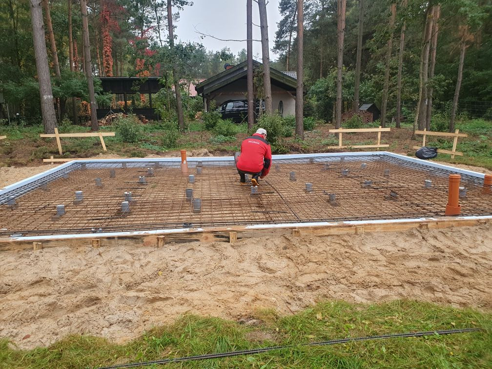 Przygotowanie zbrojenia płyty fundamentowej pod budowę domu na zalesionej działce, widoczne szalunki, siatka zbrojeniowa, dystanse i pionowe rury kanalizacyjne.
