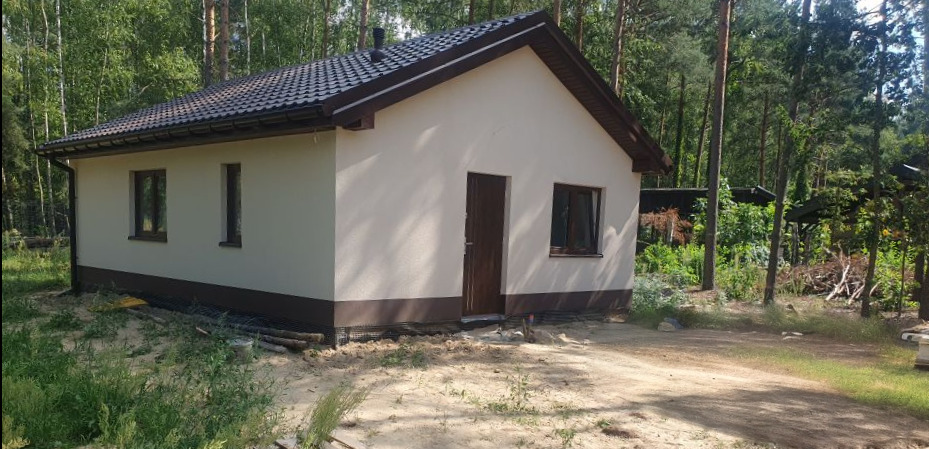 Gotowy, parterowy dom z jasną elewacją, brązową stolarką okienną i ciemnym dachem, otoczony lasem i piaskiem.