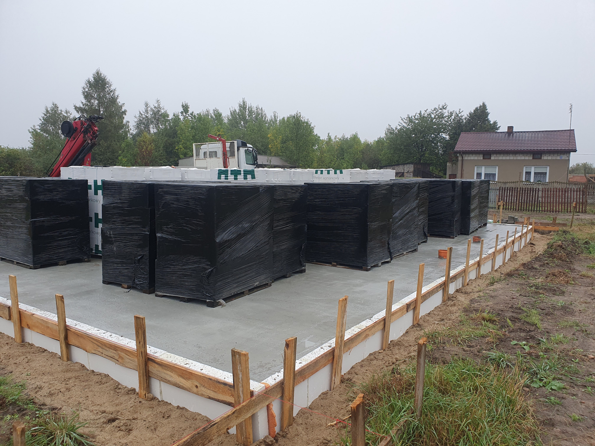 Przygotowany fundament pod budowę domu z paletami bloczków betonowych na wylewce, widoczne szalunki i dźwig w tle.