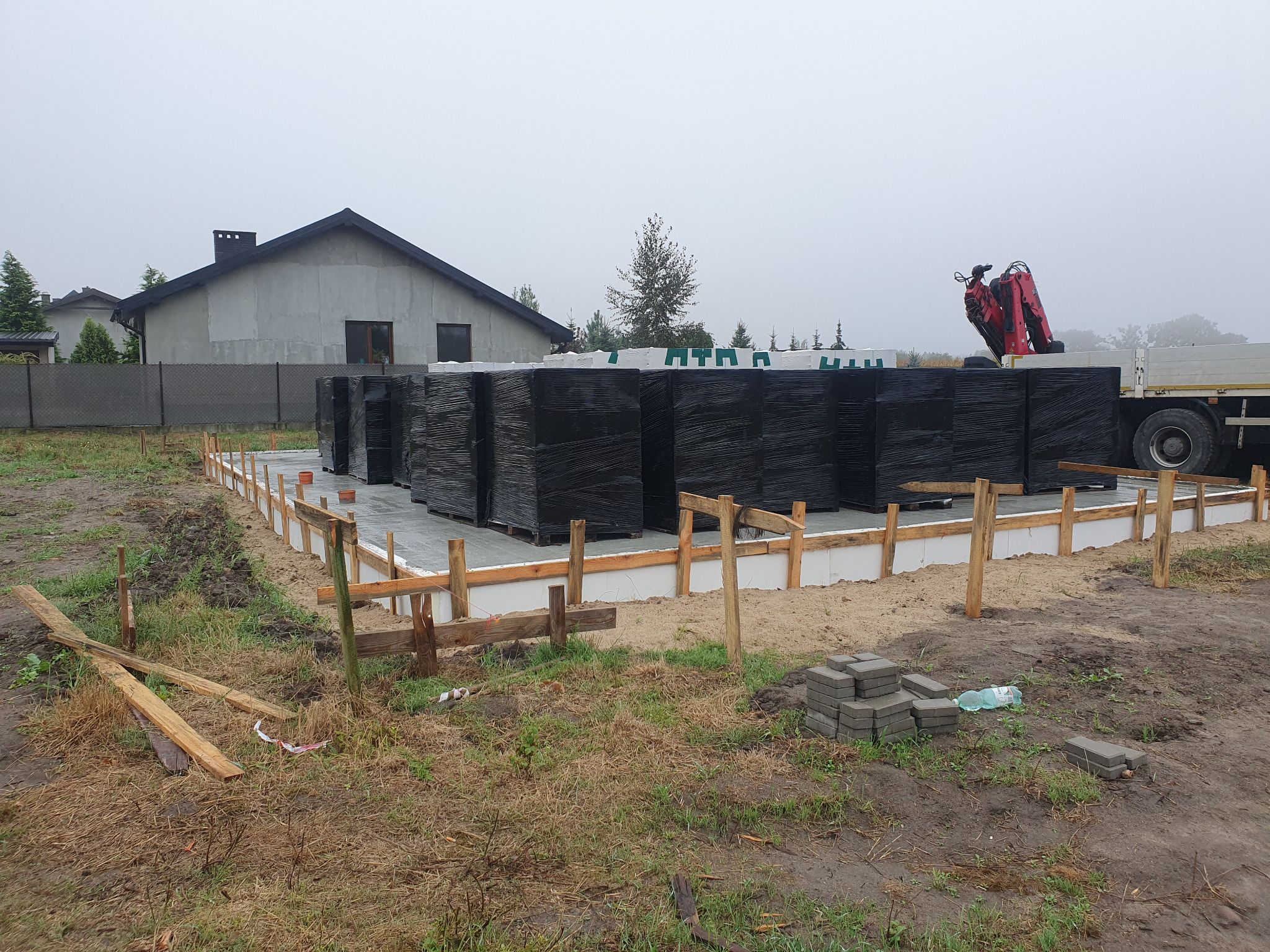 Fundament domu jednorodzinnego z widocznym szalunkiem i izolacją, przygotowany pod murowanie ścian, z paletami materiałów budowlanych na posesji i dźwigiem w tle.