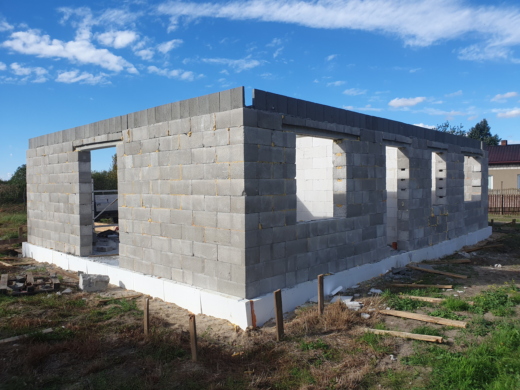 Surowy stan domu z betonu komórkowego z widocznymi otworami okiennymi i drzwiowymi, na fundamencie izolowanym styropianem, w tle błękitne niebo z chmurami.