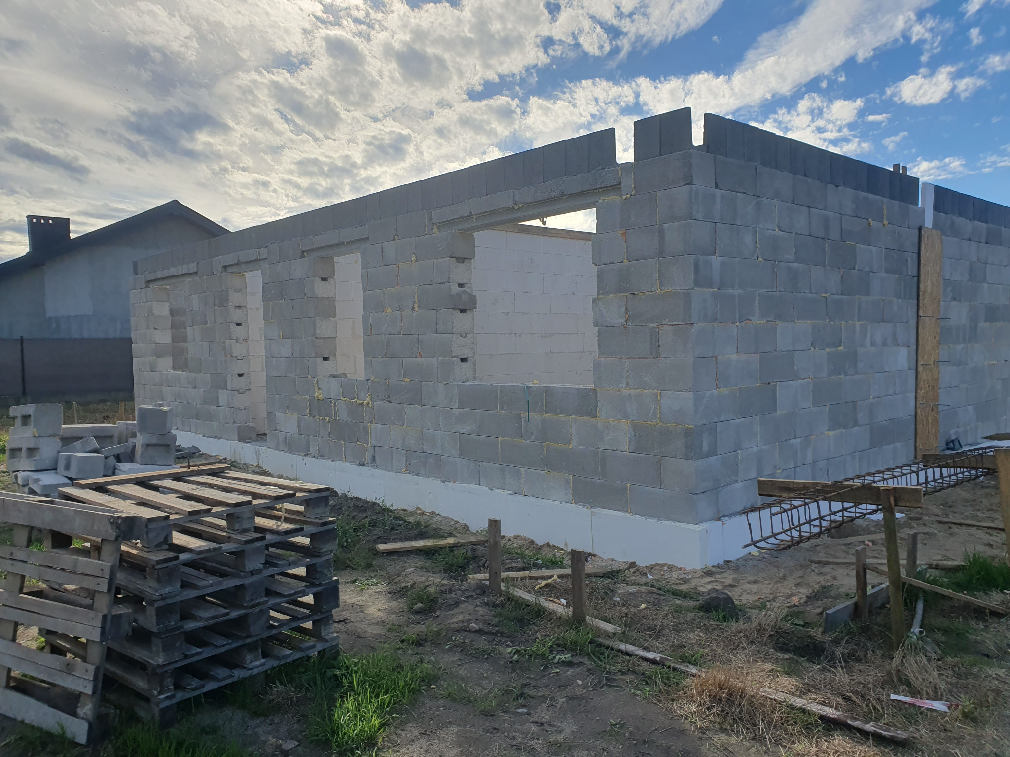 Ściany parterowego domu w trakcie budowy, wzniesione z szarych bloczków, widoczne otwory okienne i drzwiowe, ocieplenie fundamentów, paleta z bloczkami i zbrojenie na pierwszym planie, pochmurne...