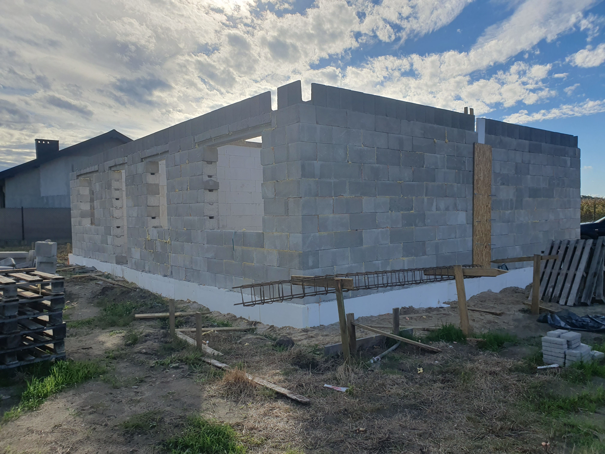 Wznoszone ściany domu z betonu komórkowego na fundamencie z izolacją, widoczne zbrojenia, palety i fragment dachu sąsiedniego budynku.