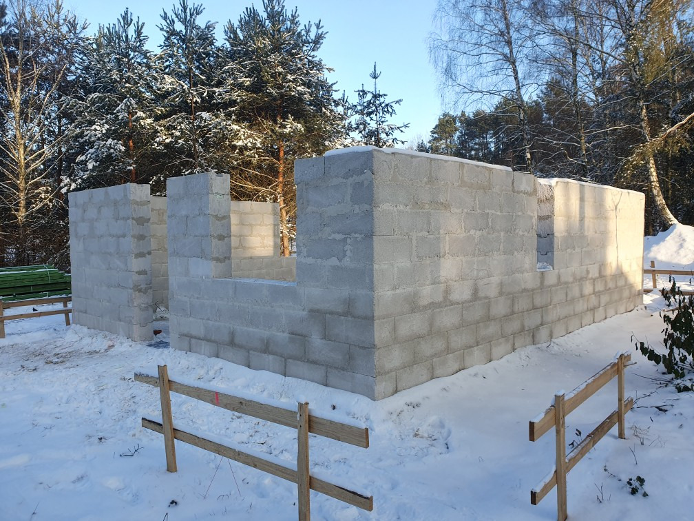 Wznoszenie ścian z bloczków betonowych w zimowej scenerii, widoczne fragmenty ścian z otworami okiennymi, ośnieżone podłoże i las w tle.
