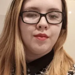 Portret kobiety w okularach, z blond włosami i bluzką w panterkę, z widocznym fragmentem koszyka wiklinowego w tle.