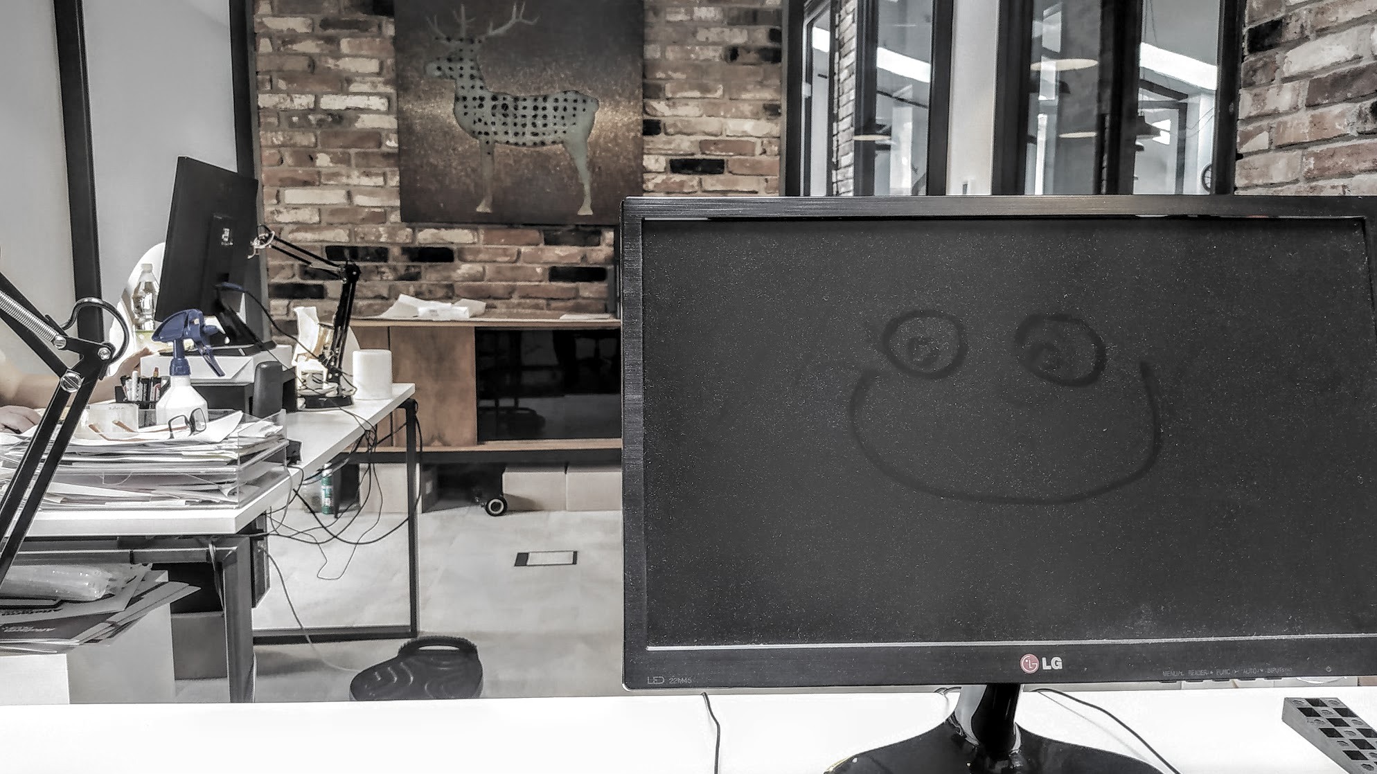 Biuro z biurkiem, stosem dokumentów, lampką i monitorem LG z narysowaną uśmiechniętą buźką na ekranie. W tle ściana z cegły i obraz z motywem jelenia.