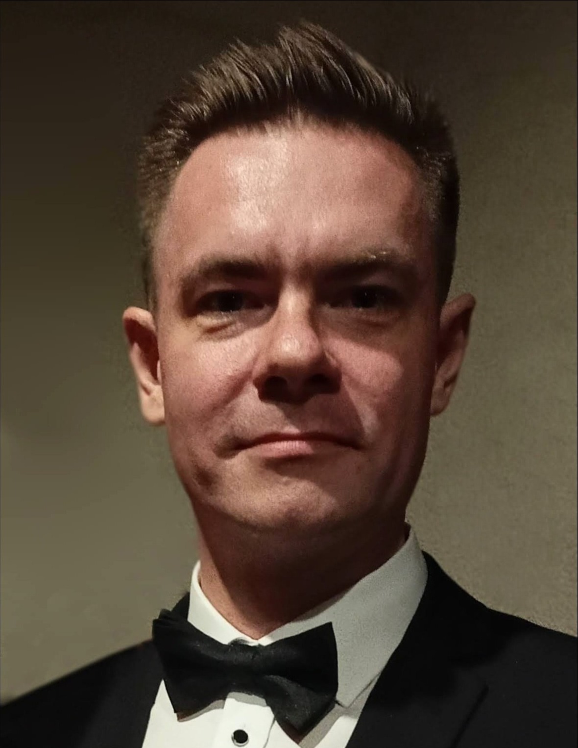 Portret mężczyzny w czarnym smokingu i muszce, z krótkimi, ciemnymi włosami zaczesanymi do tyłu, na neutralnym tle.