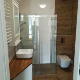 Nowoczesna łazienka z kabiną prysznicową, umywalką nablatową na drewnianym blacie, wiszącą toaletą i grzejnikiem drabinkowym.