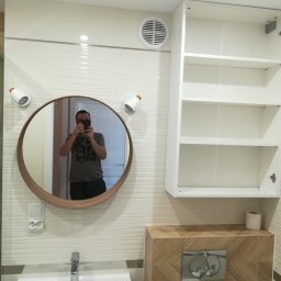 krystian moskała - Nowoczesna łazienka z okrągłym lustrem z drewnianą ramą, białymi płytkami z delikatną fakturą, białą szafką wiszącą i umywalką z chromowaną baterią. W lustrze odbija się osoba robiąca zdjęcie.