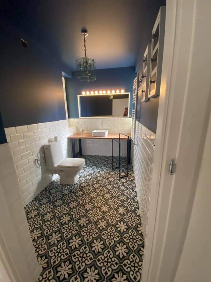 Wąska łazienka w odcieniach bieli i granatu, z dekoracyjną, wzorzystą podłogą, białymi płytkami na ścianach do połowy wysokości, drewnianym blatem pod umywalką i lustrem z oświetleniem oraz wiszącą...