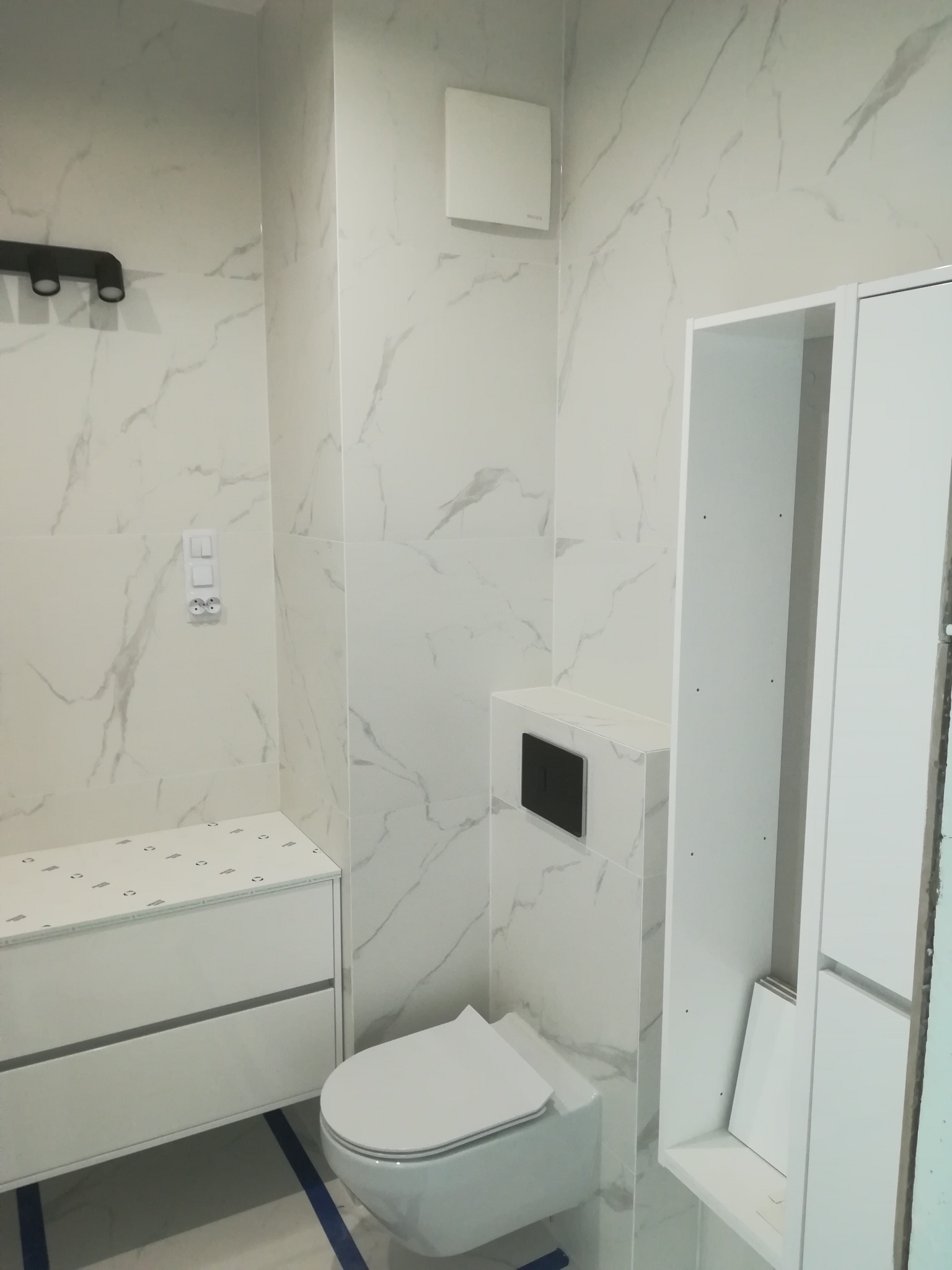 Nowoczesna łazienka w trakcie wykańczania: białe płytki imitujące marmur, podwieszana toaleta z czarnym przyciskiem, biała szafka z umywalką i wisząca szafka, zabezpieczona podłoga.