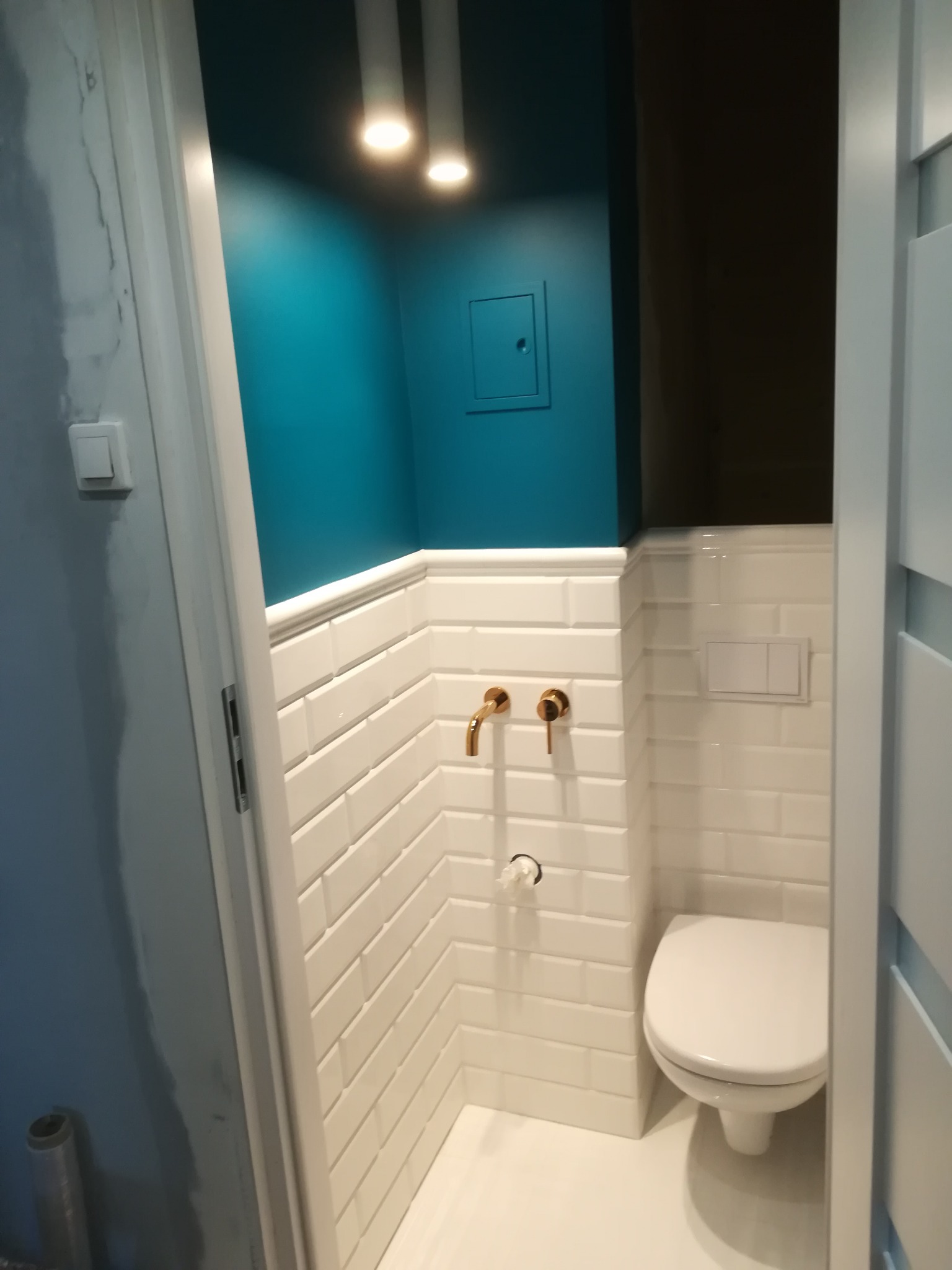 Niewielka łazienka po remoncie: białe, prostokątne płytki na ścianach do połowy wysokości, turkusowa ściana powyżej, złote krany, biała toaleta, oświetlenie punktowe.