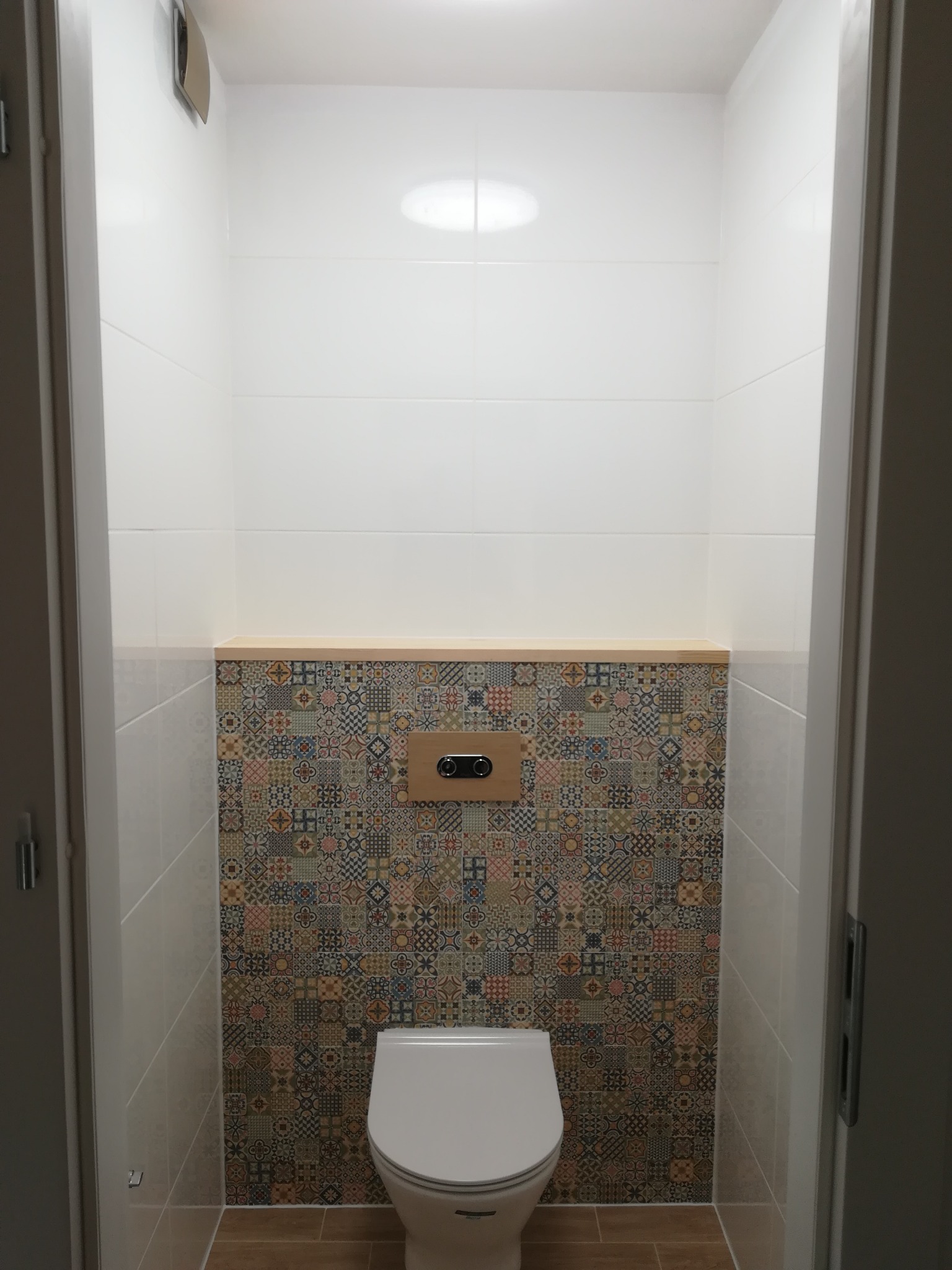 Wnętrze małej łazienki po remoncie, z białymi płytkami ceramicznymi na górze i wzorzystymi płytkami patchworkowymi poniżej, drewnianą półką i podwieszaną toaletą.