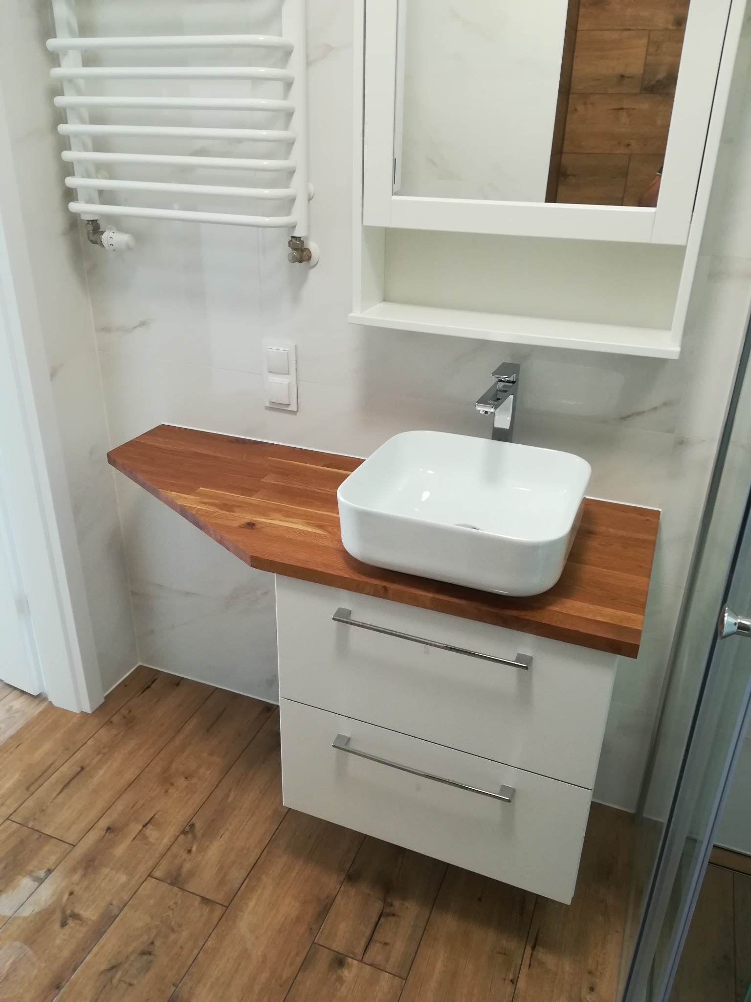 Nowoczesna łazienka z białą umywalką nablatową na drewnianym blacie, białą szafką z dwiema szufladami, grzejnikiem drabinkowym oraz marmurowymi płytkami na ścianie.