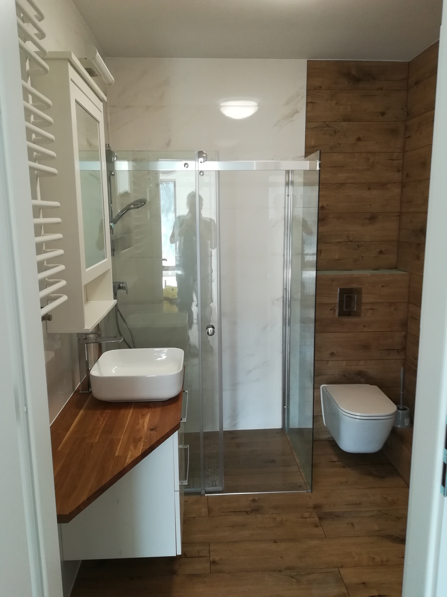 Nowoczesna łazienka z kabiną prysznicową, umywalką nablatową na drewnianym blacie, wiszącą toaletą i grzejnikiem drabinkowym.