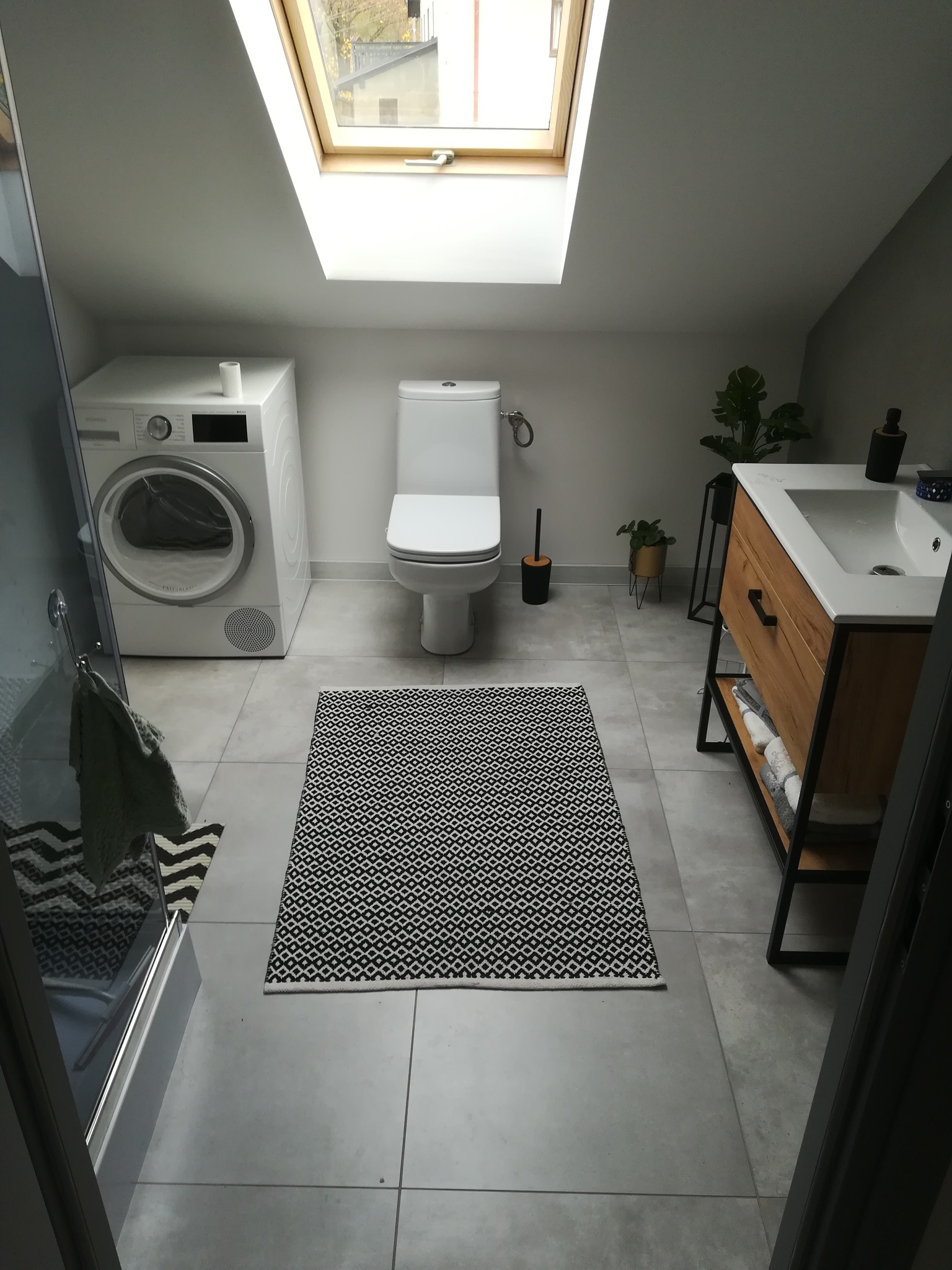 Jasna łazienka na poddaszu z oknem dachowym, szarą glazurą podłogową, białą pralką, toaletą oraz drewnianą szafką z umywalką, dywanik z geometrycznym wzorem.