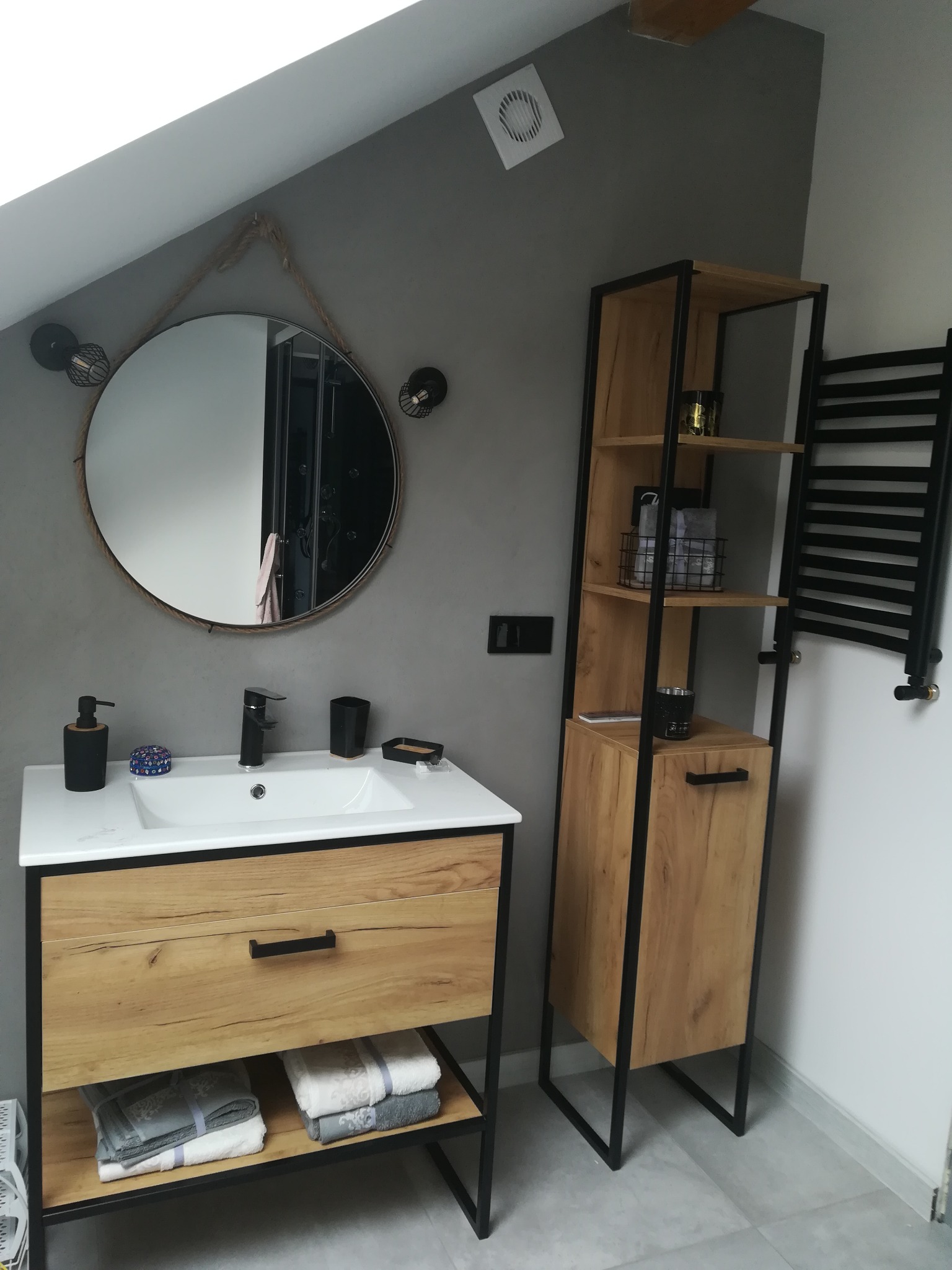 Nowoczesna łazienka na poddaszu z umywalką nablatową, okrągłym lustrem w ramie z liny, drewnianą szafką z czarnymi elementami i grzejnikiem drabinkowym.