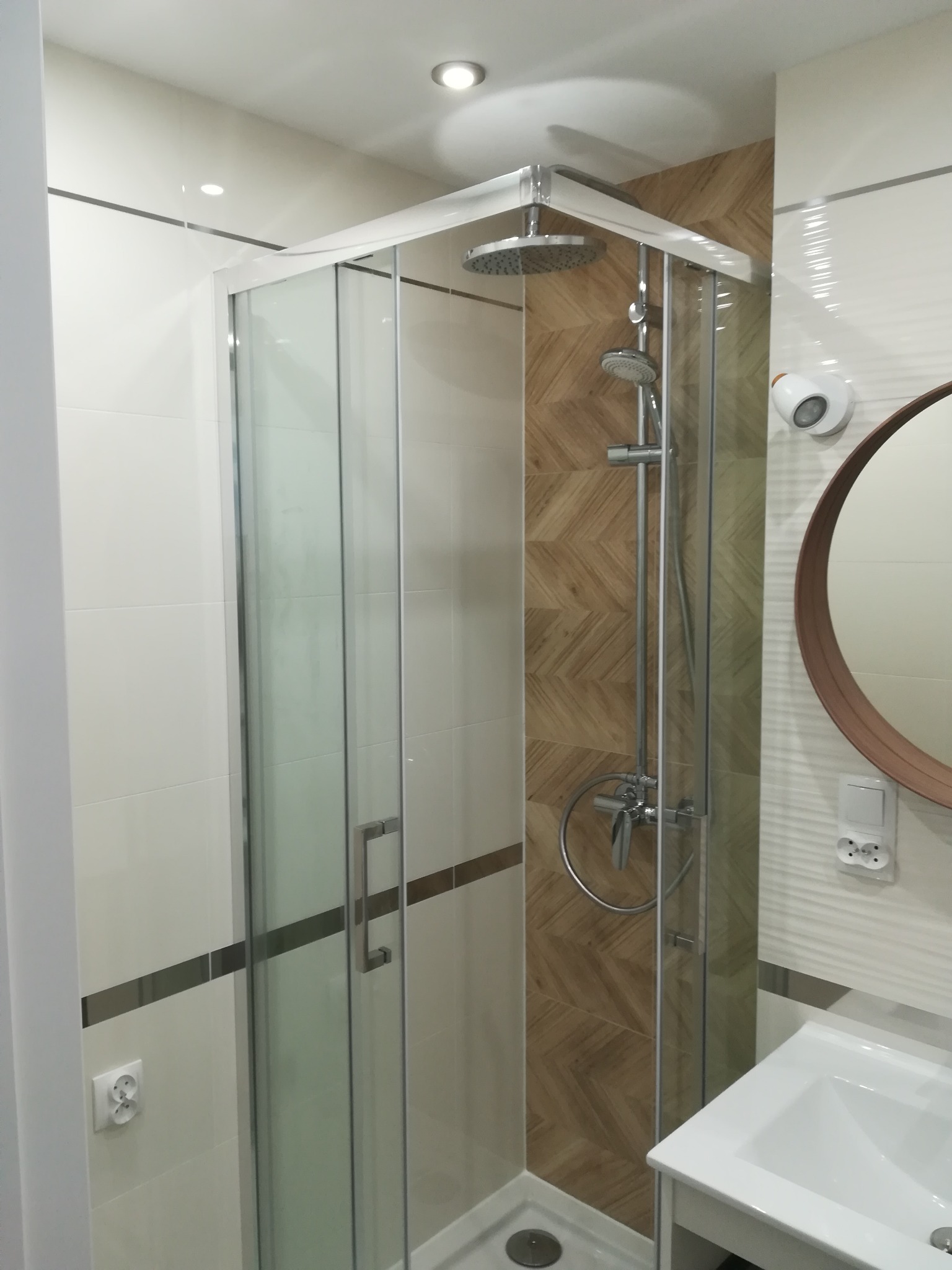 Nowoczesna kabina prysznicowa z deszczownicą, wykończona płytkami w kolorze drewna ułożonymi w jodełkę, okrągłe lustro z drewnianą ramą, biała umywalka i gniazdko elektryczne na ścianie.